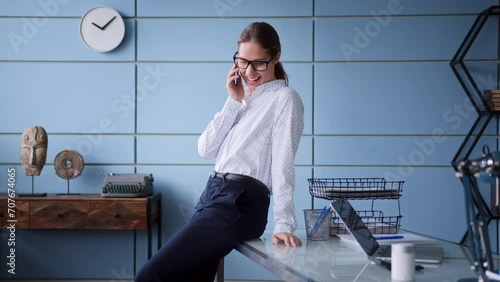 young woman in eyeglasses and a white shirt stands near table in office, chats using mobile phone, smiling and laughing, looks confident, successful and satisfied
