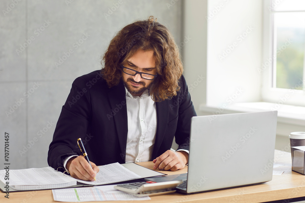 Young businessman, auditor, bookkeeper or financial accountant working in his office. Serious busy long haired man in glasses sitting at desk, doing business paperwork and using laptop computer Young businessman, auditor, bookkeeper or financial accountant working in his office. Serious busy long haired man in glasses sitting at desk, doing business paperwork and using laptop computer