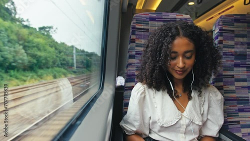 Front view of young businesswoman commuting sitting by window on moving train working on laptop and listening to music or podcast on earphones - shot in slow motion photo