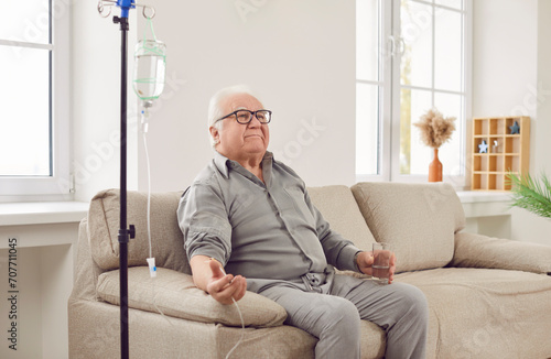 Senior man sitting on the couch at home or in clinic while receiving IV drip infusion and vitamin therapy in his blood. Smiling elderly male person in casual clothes receiving injection therapy.
