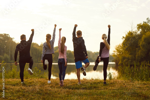 Group of friends doing fitness exercises during outdoor sports workout in nature. View from behind five people standing on one foot on withered autumn grass of river bank and raising hands up to sky
