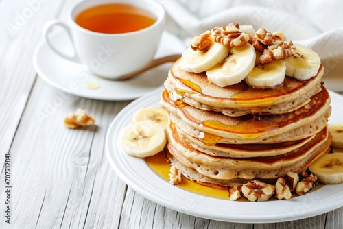 A white plate topped with banana and walnut pancakes, a cup of tea on the side, all served on a white wooden table.
