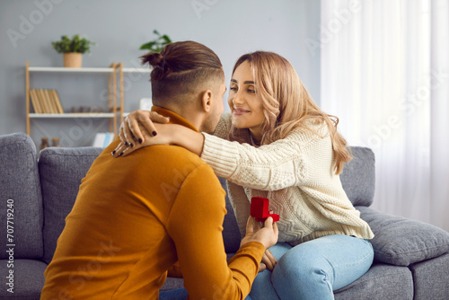 Young man giving open box with engagement ring to his happy girlfriend. Affectionate couple hugging each other while sitting on sofa at home. Marriage proposal, love, wedding concept © Studio Romantic