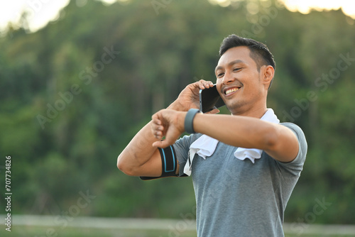 Smiling millennial man in sports clothes talking on mobile phone and checking time on watch