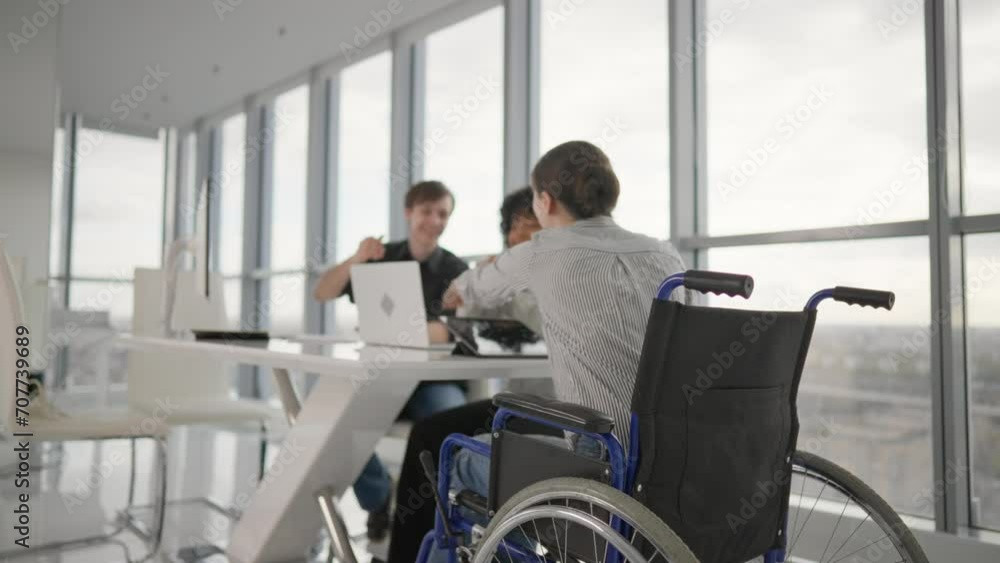 Friendly colleagues working on project in modern office together sitting at desk. Woman in wheelchair with physical disability on meeting with caucasian man african american female. Teamwork, discuss. Friendly colleagues working on project in modern office together sitting at desk. Woman in wheelchair with physical disability on meeting with caucasian man african american female. Teamwork, discuss.