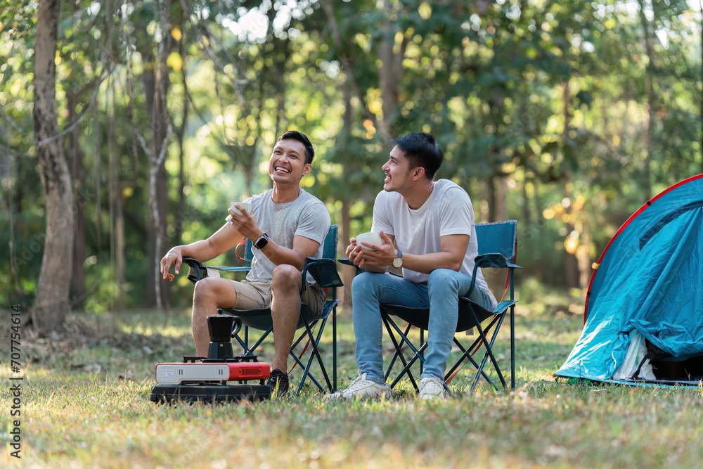 Gay LGBTQIA couple sitting on picnic chair drinking tea and coffee while camping on vacation holiday Gay LGBTQIA couple sitting on picnic chair drinking tea and coffee while camping on vacation holiday