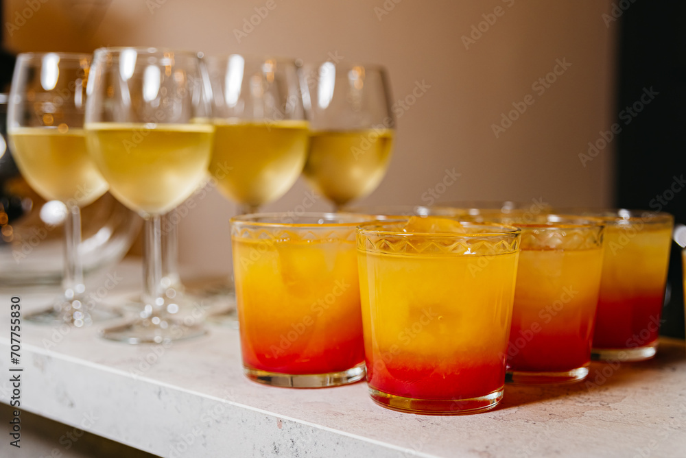 Tequila Sunrise and White Wine Selection. An array of Tequila Sunrise cocktails with gradient orange hues and white wine in stemmed glasses, ready for a party. Tequila Sunrise and White Wine Selection. An array of Tequila Sunrise cocktails with gradient orange hues and white wine in stemmed glasses, ready for a party.