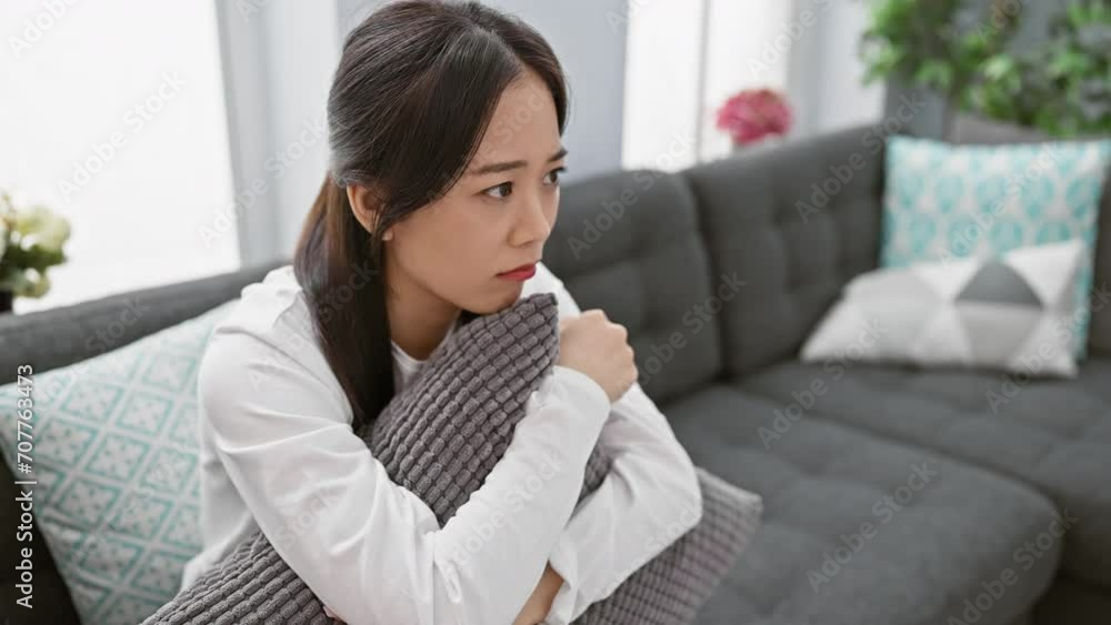 Pensive asian woman sitting indoors on couch hugging pillow, feeling contemplative Pensive asian woman sitting indoors on couch hugging pillow, feeling contemplative