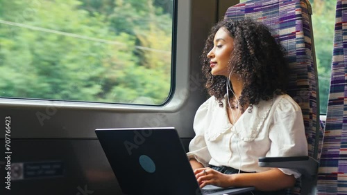 Young businesswoman commuting sitting on moving train passing through city working on laptop and listening to music or podcast on earphones looking out of window - shot in slow motion