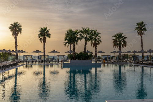 Sunrise over the pool and sunbeds. On the background of the sea coast. Big palm trees.