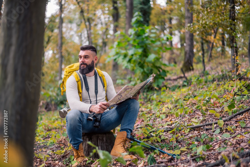 During his woodland hike, a hipster enjoys a rest, sitting on a tree stump while consulting a paper map to plan his route."