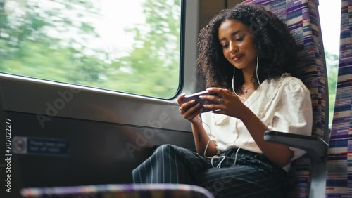 Young businesswoman commuting sitting by window on moving train streaming film or show to mobile phone wearing earphones - shot in slow motion photo