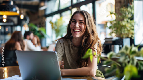 Young woman seated and smiling at the camera  with a laptop in front of her and a blurred office environment in the background