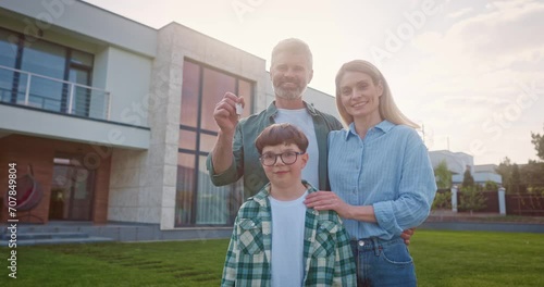 Happy family of three standing outside near big house. Smiling family of four at background of yard. Father holding keys of new house. Young family buying new appartment. Satisfied parents and kid. photo