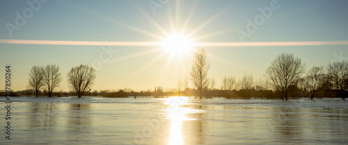 Sunset over high water level of river IJssel with vast panorama landscape and trees underwater on the horizon showing big sun flare across the wide angle view