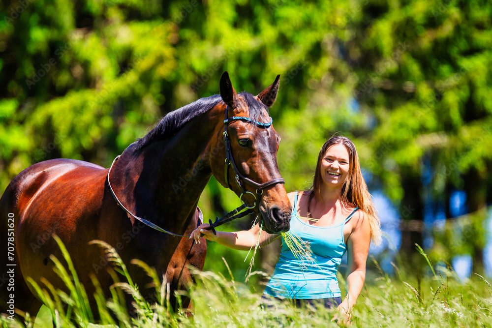 Obraz premium A young woman rider with long brunette hair stands with her horse on a high summer meadow in the bright sunshine.