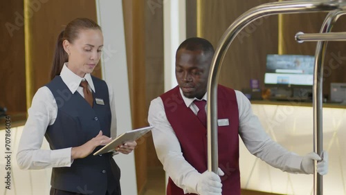 Female hotel manager using digital tablet and explaining job task to young African American porter listening to her and walking away with luggage cart photo