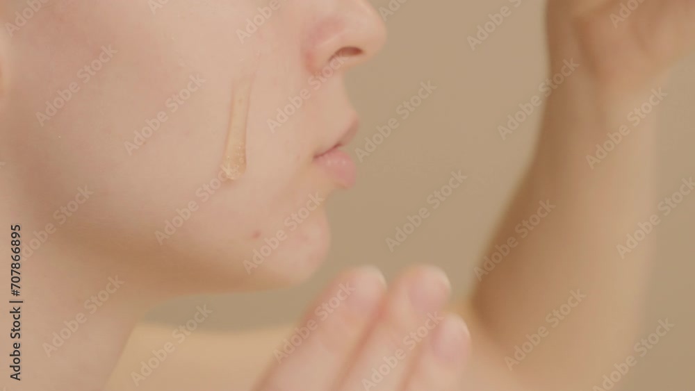 Extreme close-up shot of face Caucasian young woman puts skin care serum on her cheek from dropper on beige background. Skin care serum advertising, copy space.