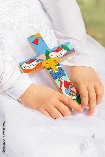 A wooden cross in the hands of a girl against the background of a white festive dress on the day of first communion, baptism. Traditions in Christianity, creed, religion and family, family traditions.