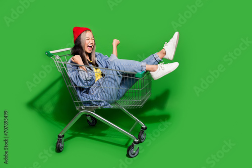 Full size photo of pretty young girl raise fists enjoy ride shopping cart dressed stylish denim outfit isolated on green color background