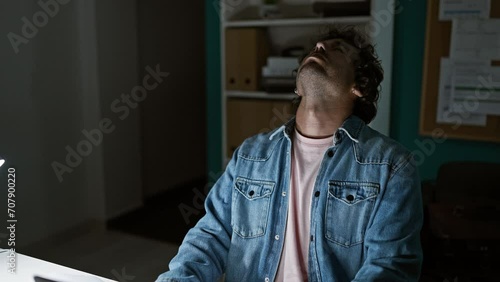 A tired hispanic man in a denim jacket expressing frustration in a dark office.
