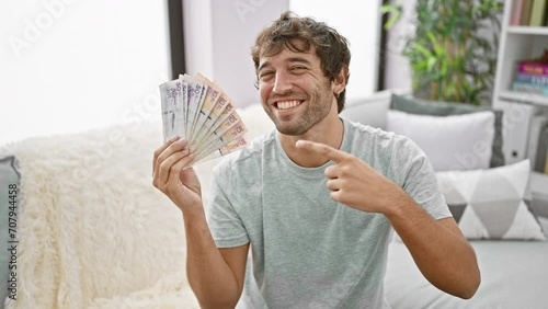 Beaming young man, holding colombian pesos at home, confidently pointing and smiling with joy photo
