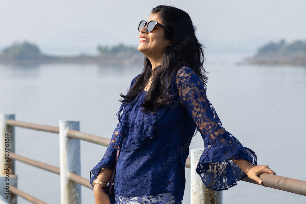 A pretty Indian woman in blue dress standing, smiling and looking at the Sun wearing sunglasses