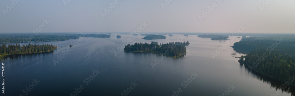 Aerial panoramic view of a large lake with many small islands. The lake is calm with small waves. The sky is filled with forest fire smoke creating a somber scene.
