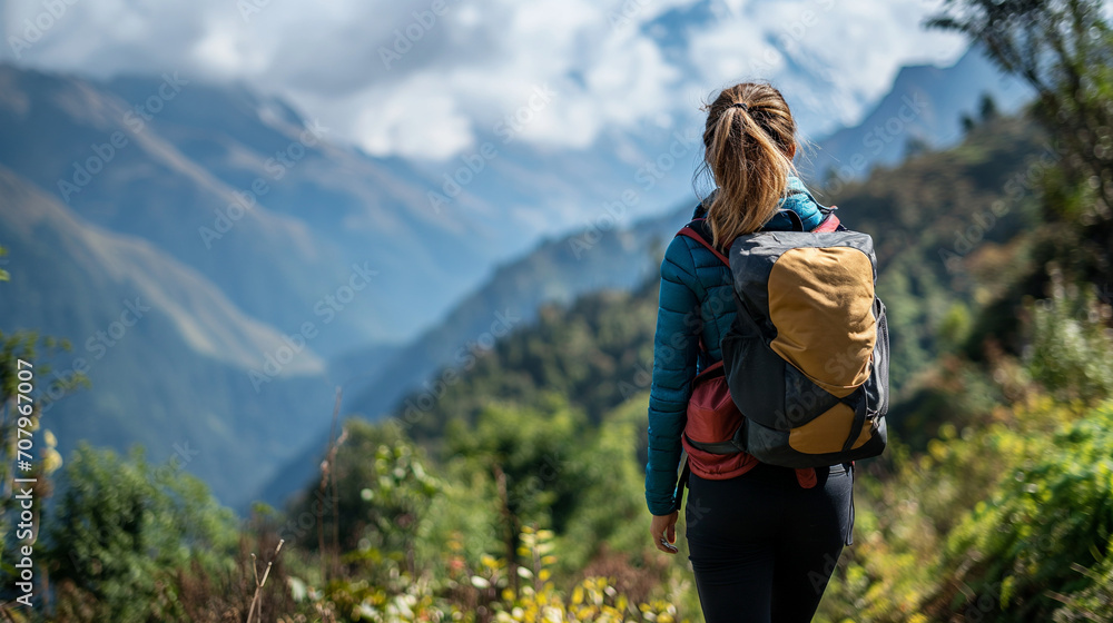 A woman with a blonde ponytail is trekking in the mountains, carrying a backpack and gazing at a distant mountain range with peaks hidden in cloud cover