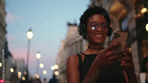 Smiling happy female checks her banking app on smartphone, ensuring her finances while enjoying music in headphones. Attractive happy black woman sending text message in social media, listen playlist photo