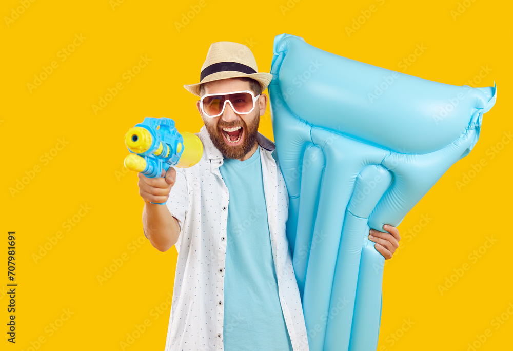 Happy cheerful man having fun on holiday. Bearded young guy in summer shirt, sun hat and glasses enjoying vacation, holding blue inflatable mattress and shooting from water gun on yellow background - obrazy, fototapety, plakaty 