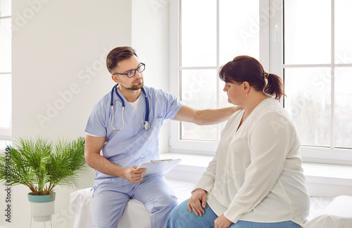 Doctor supporting sad patient. Man who works as physician at clinic is sitting on medical bed together with sick fat overweight woman, holding hand on her shoulder and expressing his compassion