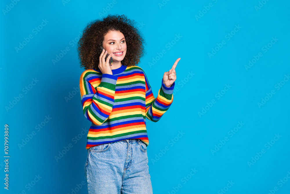Photo portrait of lovely young lady talk phone point empty space wear trendy rainbow print garment isolated on blue color background Photo portrait of lovely young lady talk phone point empty space wear trendy rainbow print garment isolated on blue color background