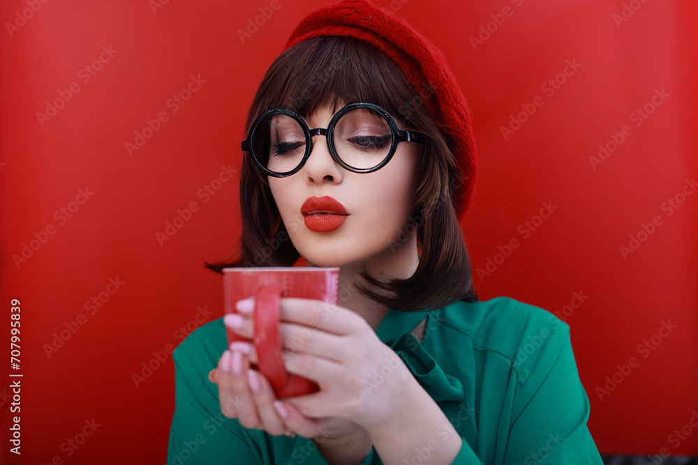 a girl with a bob hairstyle is holding a disposable glass of hot drink in her hands. the girl is wearing a shirt with a bow and a red biret. the background is red a girl with a bob hairstyle is holding a disposable glass of hot drink in her hands. the girl is wearing a shirt with a bow and a red biret. the background is red