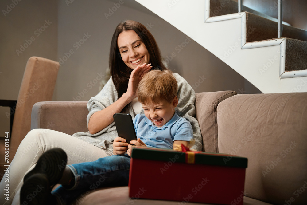 Laughing boy using smartphone with her mother on sofa celebrating Christmas Laughing boy using smartphone with her mother on sofa celebrating Christmas