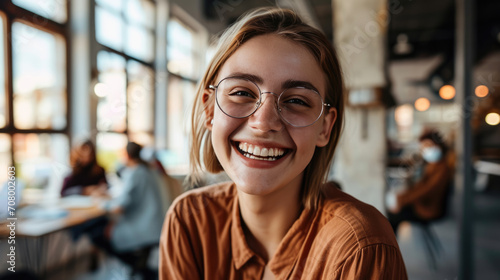 Joyful young woman with glasses is smiling broadly at the camera with a blurred office background