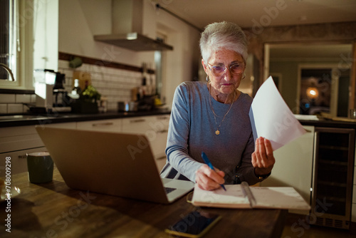 Senior Woman Working on Finances with Laptop and Papers in Kitchen at Night photo