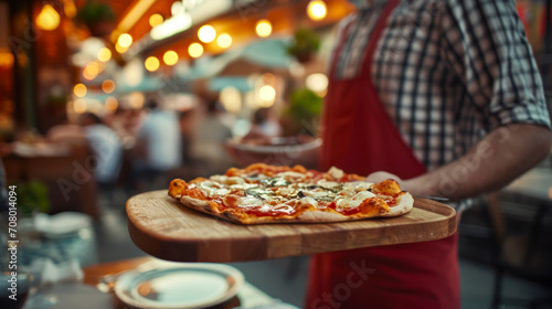 Local pizzeria, waiter carrying pizza on wooden tray to customers in old pizzeria on Italian street, selective focus on pizza, lunch concept with copy space