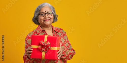 Happy Indian woman smiling senior gray-haired old lady holds a wrapped present box with gift ribbon bow on isolated on yellow colour background, studio portrait. Copy paste empty place for text