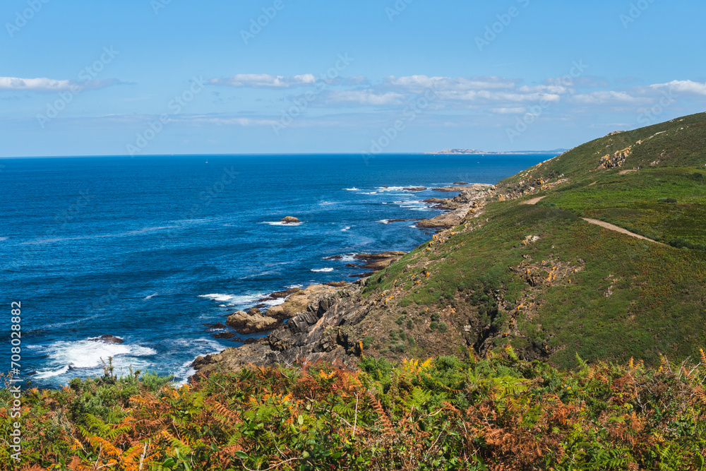Summer morning at Ons Island's western edge. Rocky cliffs adorned with lush greenery plunge into calm blue sea, ferns in the foreground. San Vicente do Mar on the distant right under a blue sky. Summer morning at Ons Island's western edge. Rocky cliffs adorned with lush greenery plunge into calm blue sea, ferns in the foreground. San Vicente do Mar on the distant right under a blue sky.