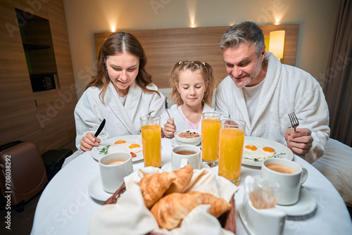 Happy caucasian family having breakfast on bed at table in hotel room