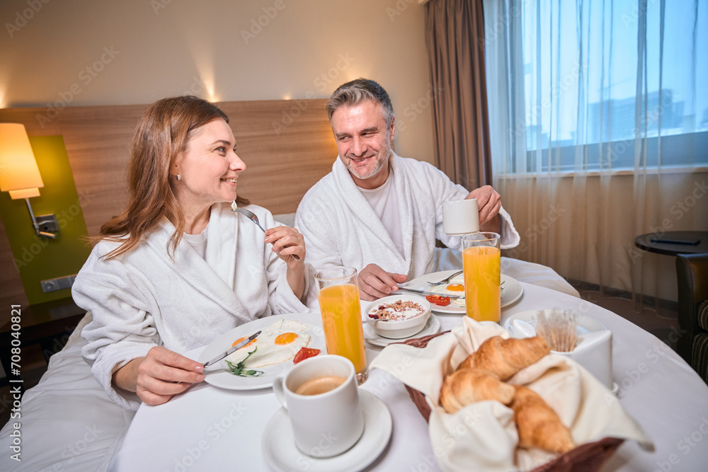 Smiling adult couple having breakfast and looking at each other in hotel room