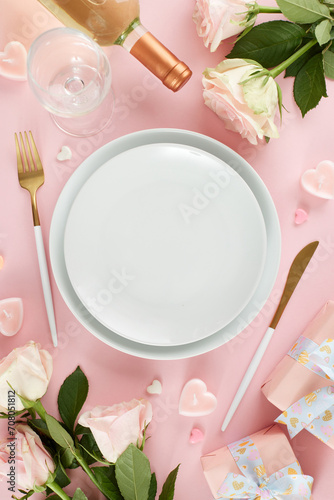 Luxurious Valentine's Day dining affair. Top view vertical photo of plates, cutlery, hearts, gift boxes, wine bottle, wine glass, roses on pastel pink background with promo zone