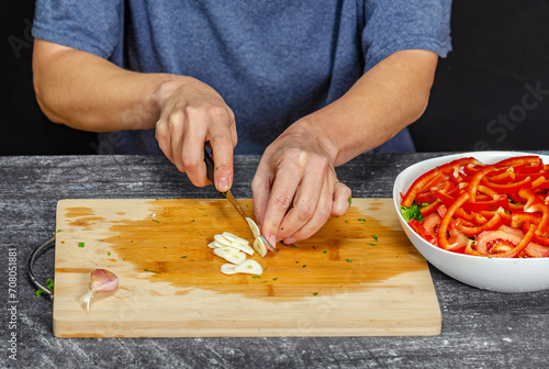 Hands of woman chopping fresh garlic on wooden board. Bowl salad on gray table, black background.