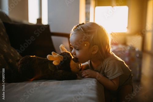A toddler kissing a plush reindeer on nose, on a sofa, in a warmly lit room.
