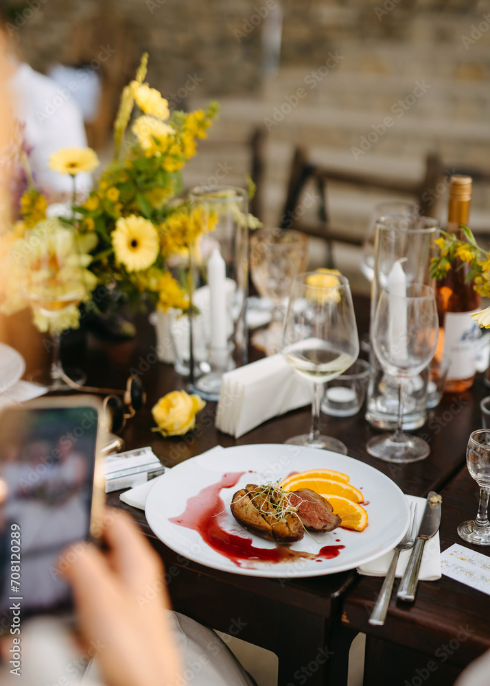 A guest captures a photo of a gourmet dish at a floral adorned table setting. A guest captures a photo of a gourmet dish at a floral adorned table setting.