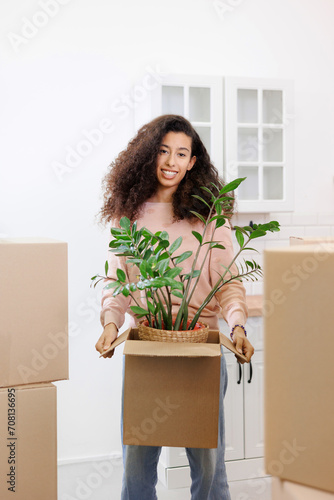 happy young African American woman is moving into new home. person stands among cardboard boxes and smiles, holding a houseplant, mortgage or rental property in his hands. mover service for moving. © MyJuly