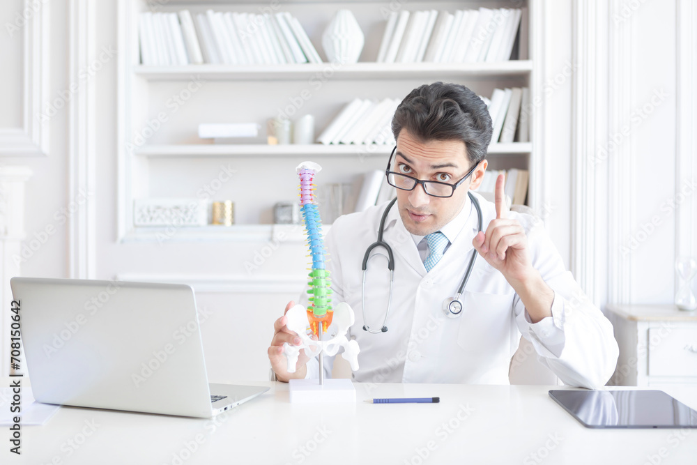 Young handsome male doctor orthopedist demonstrating the problem on spine bone model on the desk in his workplace Young handsome male doctor orthopedist demonstrating the problem on spine bone model on the desk in his workplace
