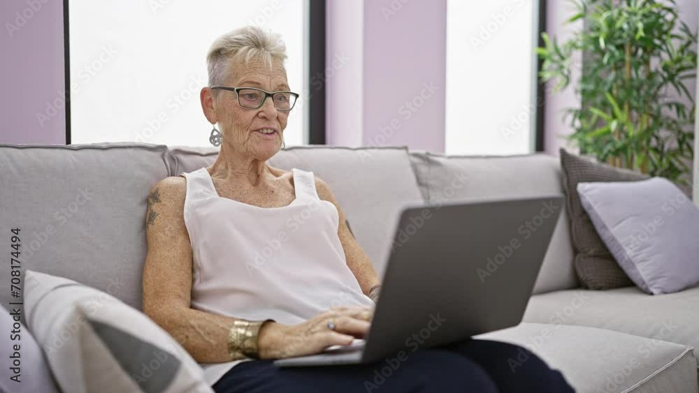 Confident grey-haired senior woman, smiling and enjoying restful moment on sofa at home, engaging with technology using laptop for relaxing video call Confident grey-haired senior woman, smiling and enjoying restful moment on sofa at home, engaging with technology using laptop for relaxing video call