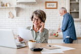 Busy european businesswoman old senior grandmother wife working, doing paperwork, dealing with documents while husband cooking dinner at home kitchen. Equality